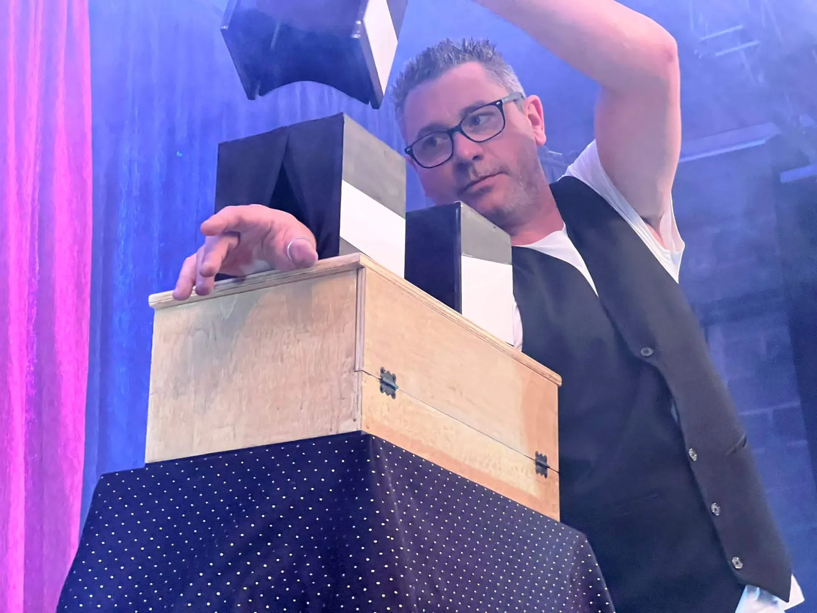 Vince sur scène, penché au-dessus d’une caisse en bois d’où jaillissent des boîtes noires et blanches, devant un rideau rose sous une lumière bleue